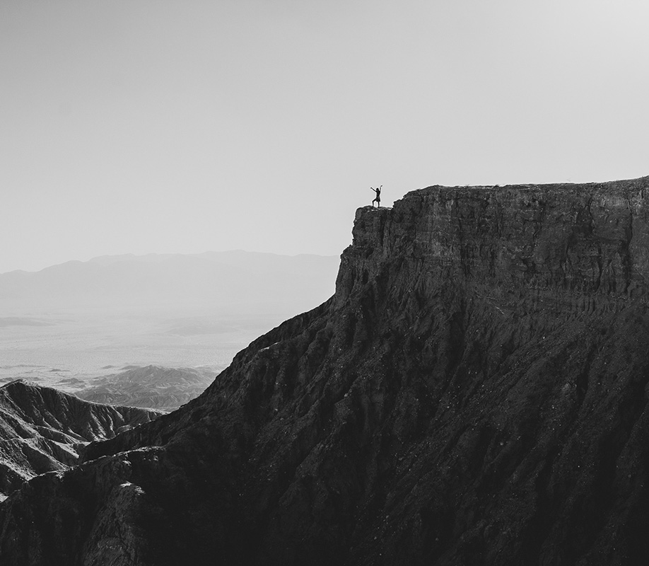 Photo en noir et blanc d'une femme brandissant les bras en l'air, en haut d'une falaise, regardant la vallée en contrebas.