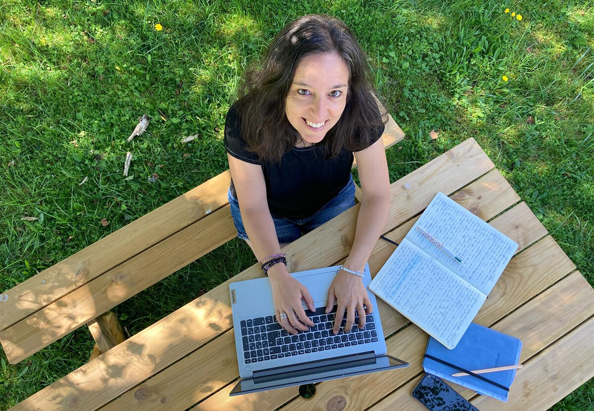 Florence Gotschaux, installée sur une table en bois, dans la verdure, en train de taper sur son ordinateur.