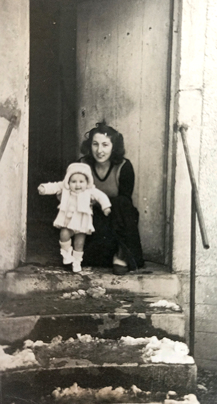 Photo en noir et blanc, datant de 1950 montrant une jeune femme tenant un bébé de quelques mois, sur le pas de la porte d'une maison, en haut de quelques marches.
